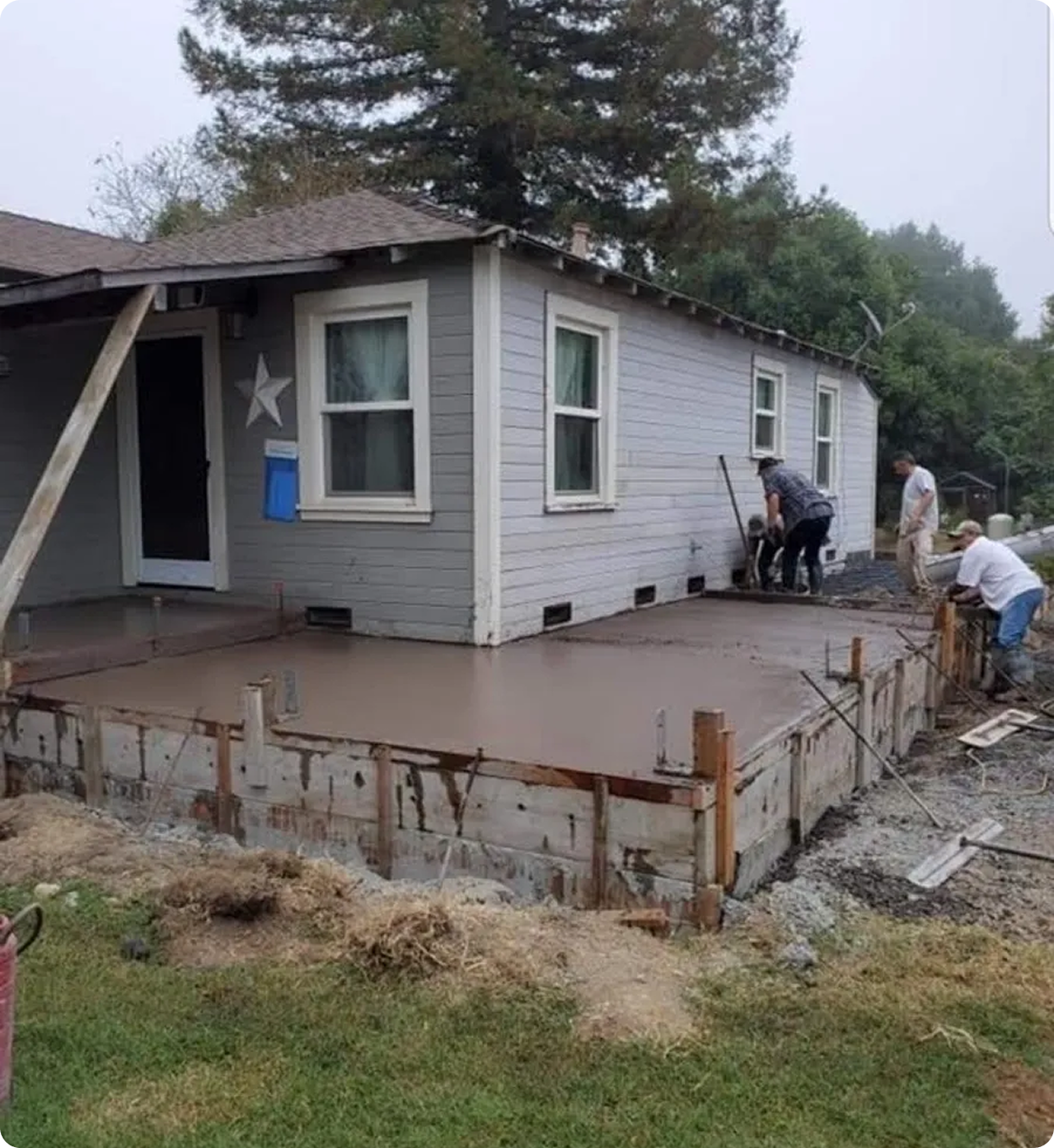 Bear State Construction Several people work on laying concrete for an extension at the back of a gray house with a small roof and two windows.