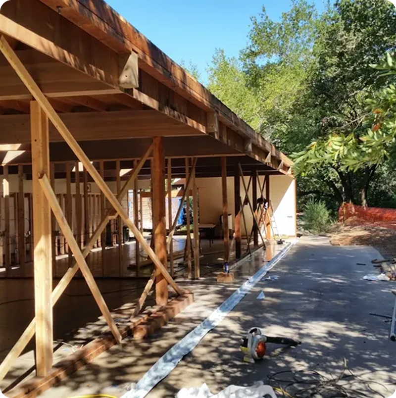 Wooden house under construction, featuring exposed beams and partial walls, with tools scattered on the ground; trees and clear sky in the background.