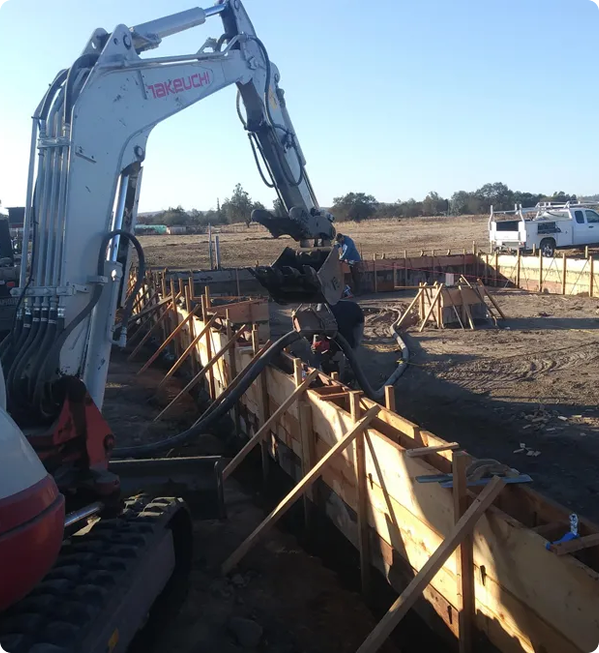 Bear State Construction Excavator pouring concrete into wooden foundation forms at a construction site, with vehicles and a barren landscape in the background.