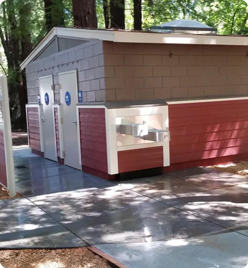 Red and gray public restroom facility with two doors and a drinking fountain outside, surrounded by trees.