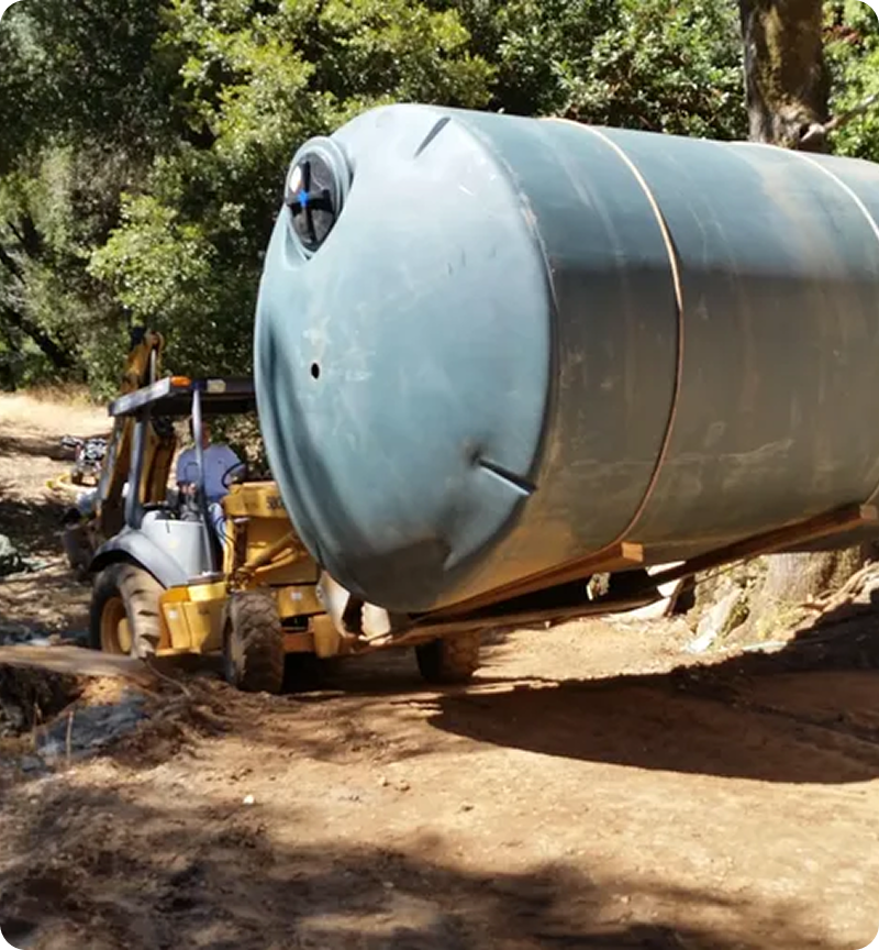 A backhoe loader moves a large cylindrical water tank through a dirt area surrounded by trees.