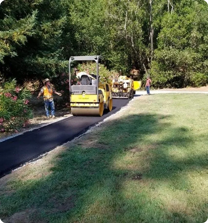 Workers paving a path with a steamroller and asphalt machine in a park setting surrounded by trees and grass.