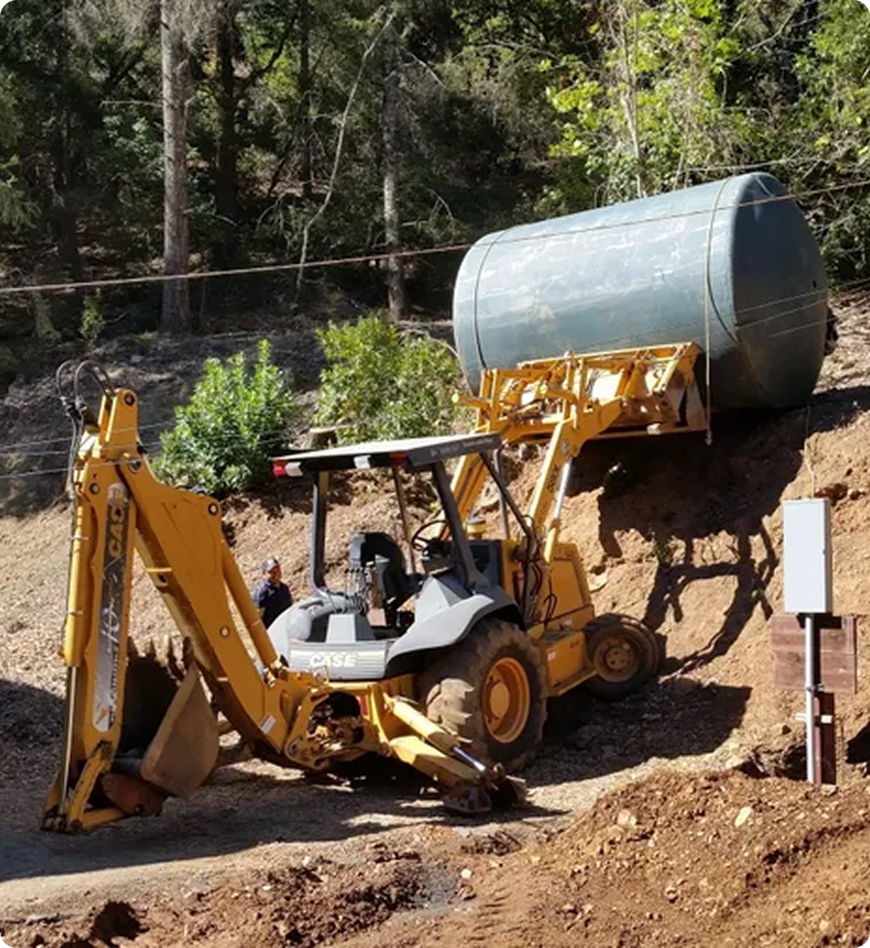 Bear State Construction Yellow backhoe loader lifting a large metal cylinder on a dirt slope, with trees in the background.